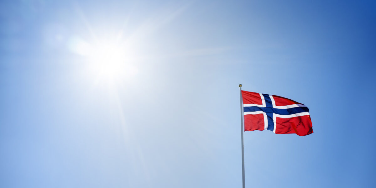 Det norske flagget på toppen av en flaggstang. Blå himmel med sol i bakgrunnen. Foto.