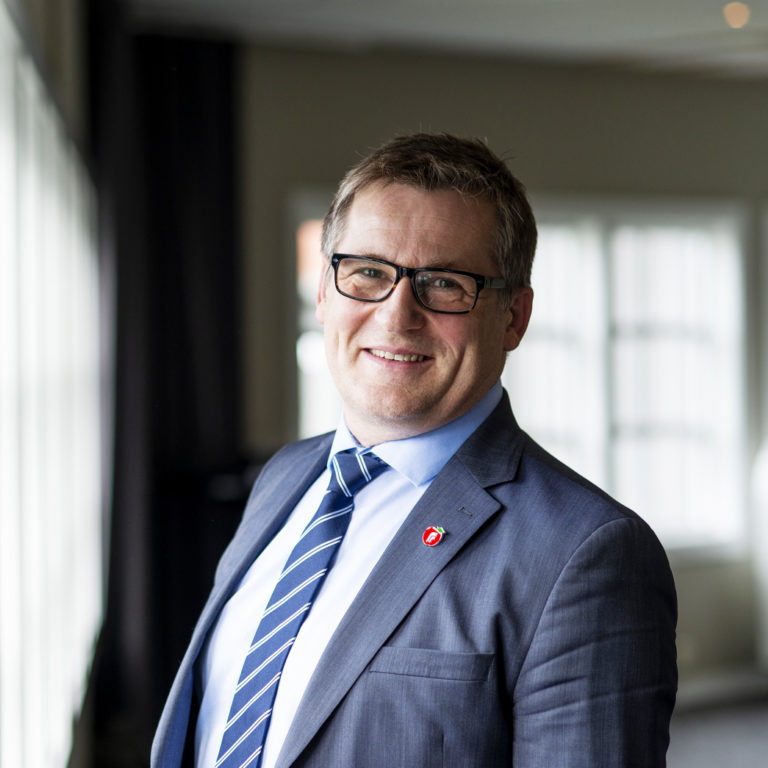 Dagfinn Henrik Olsen, i blå dress, smilende foran et vindu. Portrett. Foto.