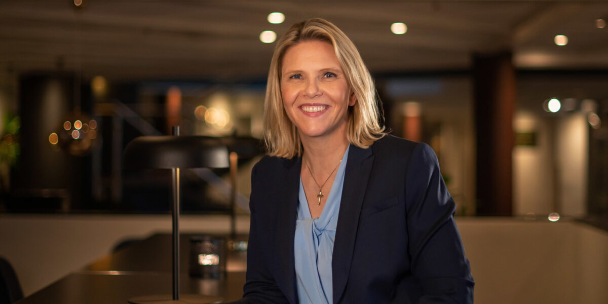 Sylvi Listhaug, smilende i blå dressjakke og lyseblå overdel. Lener seg svakt fremover mot et bord. Uskarp bakgrunn. Foto.