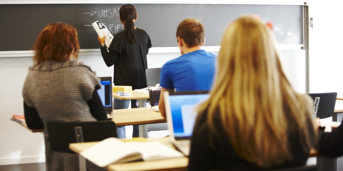 Tre elever med ryggen til i klasserom. Lærer skriver på tavle. Foto. Tre elever med ryggen til i klasserom. Lærer skriver på tavle. Foto.